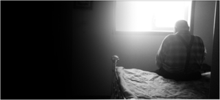 An elderly man sitting on a bed and looking out a window